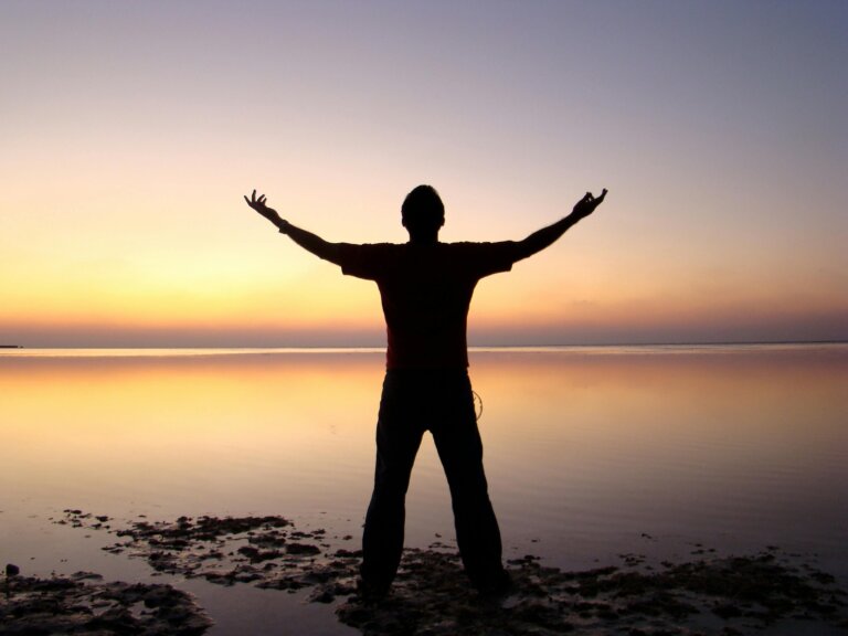 A man standing with one hand raised toward the sunset, symbolizing hope, strength, and freedom after overcoming challenges.